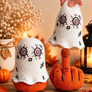 May include: Two crocheted figures featuring white ghost covers with embroidered red spiral eyes and black stitching details, placed over orange crocheted bases. One figure is shaped like a hand making an obscene gesture.