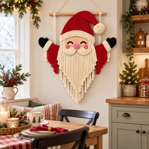 May include: A macrame wall hanging shaped like Santa Claus, featuring a red hat with a cream pom-pom, a long cream beard, pink cheeks, and black mittens. The piece is suspended from a wooden dowel with white cord, showcasing intricate knotting.