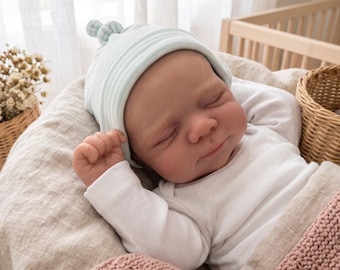 Puede incluir: Una muñeca realista de recién nacido con los ojos cerrados y una sonrisa sutil, que lleva un body blanco de manga larga y un gorro con nudo a rayas de color verde menta claro y blanco, sobre una tela beige suave.