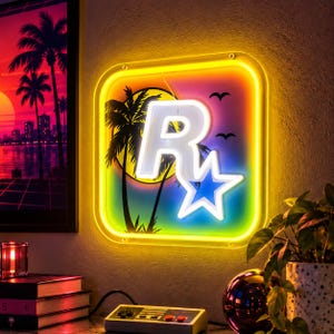 May include: A square neon sign featuring a white Rockstar Games logo with a star, set against a vibrant sunset background with palm tree silhouettes and birds. The sign is framed by a bright yellow neon border, emitting a warm glow.