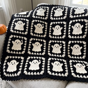 Puede incluir: Una manta de ganchillo hecha a mano con una cuadrícula de cuadrados negros, cada uno con un motivo de fantasma color crema. El borde de cada cuadrado está decorado con pequeños corazones de ganchillo blancos. Borde negro sólido.