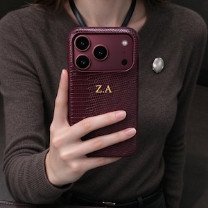 May include: A deep burgundy smartphone case with a textured lizard-skin pattern and a matte finish. The case features a prominent camera module cutout and the gold-colored initials Z.A embossed in the center of the back panel.