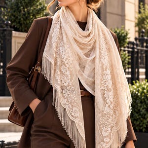May include: A cream-colored triangular lace scarf featuring an intricate floral pattern and long fringe trim along the edges. The scarf is draped over a dark brown blazer and matching trousers, paired with a brown leather belt and handbag.