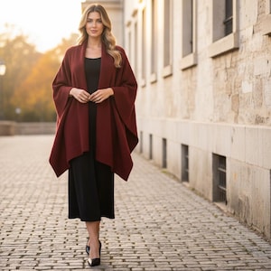 May include: A deep burgundy open-front knit poncho worn over a simple black sleeveless dress. The poncho features a wide shawl collar, loose draped sleeves, and a hem that falls below the hips, creating a layered silhouette with clean lines.