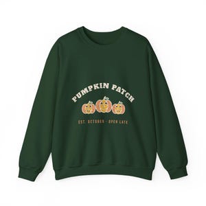 May include: A dark forest green crewneck sweatshirt featuring a graphic of three orange pumpkins with carved faces. The text PUMPKIN PATCH is arched above the pumpkins, and the text EST. OCTOBER - OPEN LATE is printed below in orange.