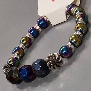 May include: A beaded bracelet featuring iridescent, multi-colored spherical beads, faceted dark blue beads, and silver-toned metal spacers. A silver-toned flower-shaped charm is included. A white tag attached to the bracelet displays the text Handmade with love.