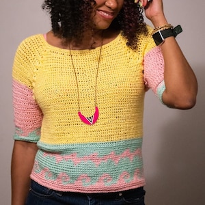 May include: A woman wearing a yellow, pink, and green crochet top with a green hat and blue denim shorts.