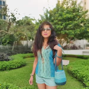 Puede incluir: Una mujer con un top de crochet azul y verde con un bolso de crochet a juego. Lleva pantalones cortos de mezclilla azul y tiene el pelo largo y oscuro. Lleva gafas y un collar.