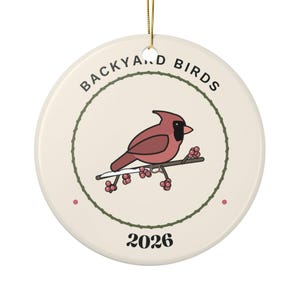 Può includere: Un ornamento rotondo in ceramica color crema con un cardinale rosso appollaiato su un ramo con bacche. Il testo BACKYARD BIRDS è stampato in nero in alto e l'anno 2026 in basso, il tutto all'interno di un cerchio verde.
