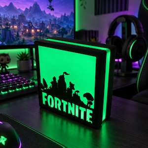 Peut inclure: Une boîte lumineuse carrée à cadre noir avec un panneau rétroéclairé vert vif. La face avant présente une silhouette noire de paysage avec des arbres et le mot FORTNITE en lettres capitales grasses au bas de la surface lumineuse.