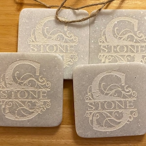 May include: Four white marble coasters with a decorative design and the word "STONE" engraved on each. The coasters are square and have a speckled surface.
