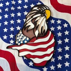 May include: A layered wooden bald eagle sculpture with a patriotic theme. The eagle is depicted with a brown body, white and black feathers, and a yellow beak. The sculpture is set against a backdrop of a blue and white star-patterned fabric and a red and white striped fabric.