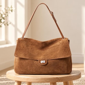 Puede incluir: Un bolso de hombro de ante marrón con una solapa rectangular, un cierre de giro de metal plateado y una correa fina ajustable sujeta con herrajes plateados. El bolso tiene una superficie suave y texturizada con base.