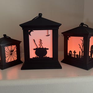 May include: Three black lanterns with orange glowing panels. The smallest features a spider web, the middle one shows a cauldron with bubbles, and the largest displays a graveyard scene with tombstones, a bare tree, and a ghost silhouette.