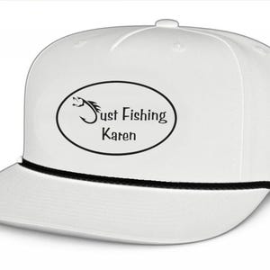 May include: A white snapback cap featuring a black rope detail across the base of the crown. The front displays an oval graphic containing a black fish hook icon and the text Just Fishing Karen in a black sans-serif font.