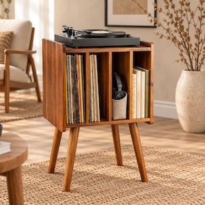 May include: A brown wooden record player stand with four tapered legs, featuring vertical storage compartments filled with vinyl records, books, and a pair of black headphones in a fabric holder, topped with a black turntable.