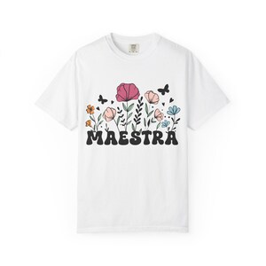 May include: A white short-sleeve t-shirt featuring a graphic print of colorful flowers, small black hearts, and butterflies above the word MAESTRA written in a bold, black, rounded font across the center of the chest area.