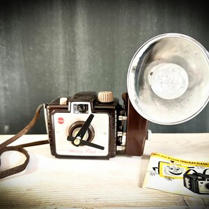 Puede incluir: Una cámara vintage Kodak Brownie Holiday Flash de color marrón y blanco con flash incorporado y una correa negra. La cámara tiene una cara blanca con un dial negro y un círculo rojo con el logotipo de Kodak. La cámara está sentada sobre una superficie de madera clara con un folleto de instrucciones amarillo.