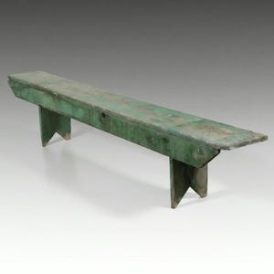 May include: A long, narrow wooden bench painted in a weathered green finish. The bench features a flat rectangular seat and two sturdy, V-cut support legs at each end. The wood surface shows signs of wear, revealing texture and grain.