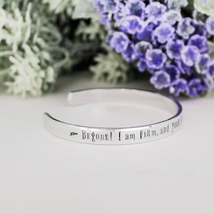 May include: A silver cuff bracelet engraved with the words "Begone! I am firm, and you shall not pass!" The open bracelet is shown against a backdrop of white and purple flowers.