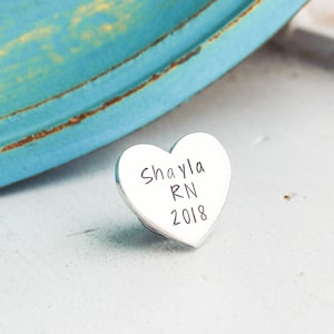 Może przedstawiać: Mały, srebrny wisiorek w kształcie serca z napisem "Shayla RN 2018" w odręcznej czcionce. Urok jest lekko pochylony, prezentując swoją wypolerowaną powierzchnię. Tło obejmuje turkusowy obiekt i białą powierzchnię.