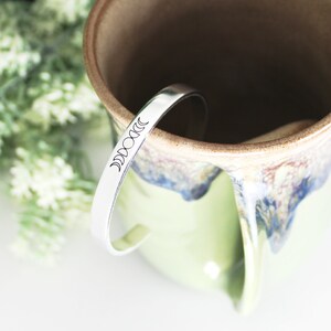 May include: A silver cuff bracelet featuring a crescent moon phase design. The bracelet is placed on a green and brown ceramic mug. The mug has a speckled glaze and a large handle. White flowers are in the background.