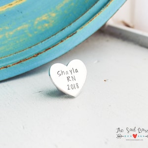 May include: A small, heart-shaped, silver-colored charm with the engraved text "Shayla RN 2018". The charm is resting on a white surface, with a blurred turquoise and yellow background.