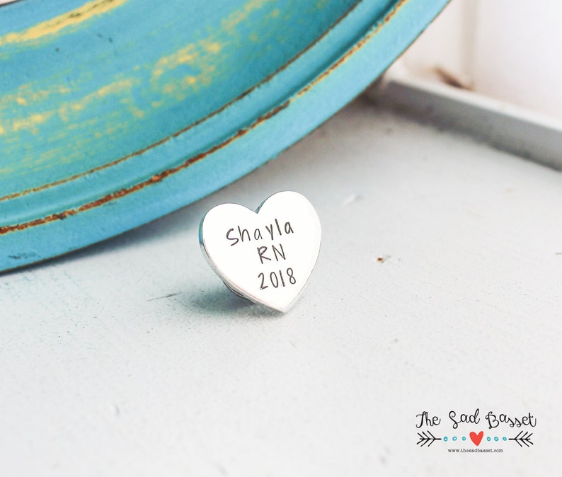 May include: A small, heart-shaped, silver-colored charm with the engraved text "Shayla RN 2018". The charm is resting on a white surface, with a blurred turquoise and yellow background.