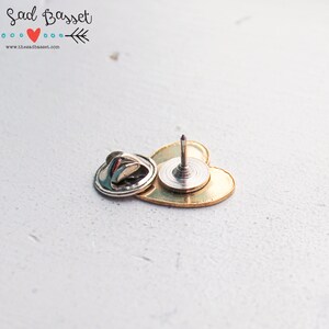 May include: A gold-colored heart-shaped pin with a silver backing and a sharp, pointed pin. The pin is designed to be attached to fabric or other materials. The text "The Sad Basset" is visible in the upper left corner.
