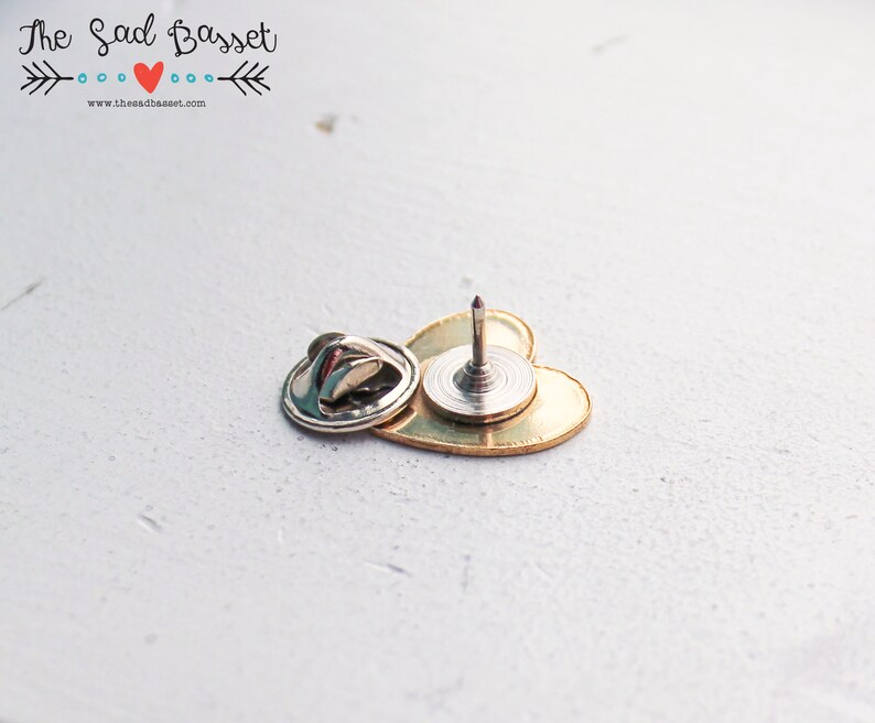 May include: A gold-colored heart-shaped pin with a silver backing and a sharp, pointed pin. The pin is designed to be attached to fabric or other materials. The text "The Sad Basset" is visible in the upper left corner.