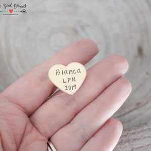 May include: A heart-shaped, gold-colored pin with the name "Bianca", the letters "LPN", and the year "2019" engraved on it. The pin is held in a hand.