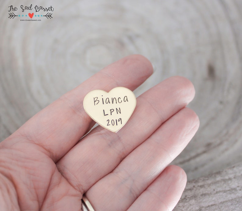 May include: A heart-shaped, gold-colored pin with the name "Bianca", the letters "LPN", and the year "2019" engraved on it. The pin is held in a hand.