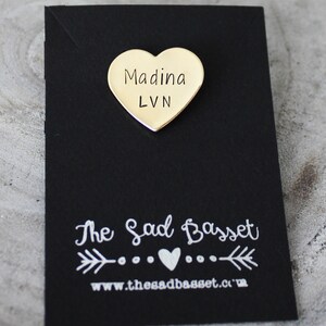 May include: A gold-colored heart-shaped pin with the name "Madina" and the letters "LVN" printed on it. The pin is on a black card with the text "The Sad Basset" and a website address.