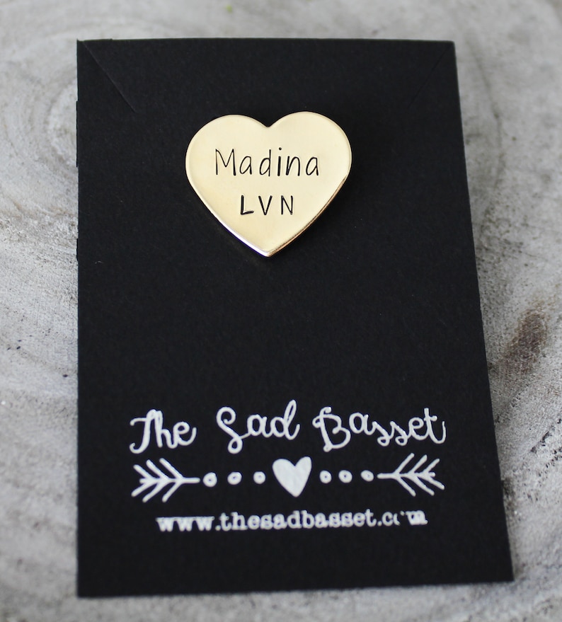 May include: A gold-colored heart-shaped pin with the name "Madina" and the letters "LVN" printed on it. The pin is on a black card with the text "The Sad Basset" and a website address.