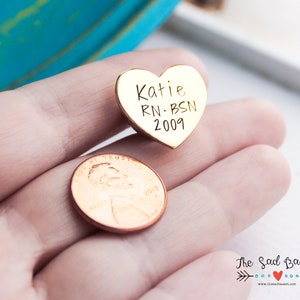 May include: A gold-colored heart-shaped pin with the engraved text "Katie RN BSN 2009". A penny is next to the pin. The pin is likely a personalized gift or keepsake.
