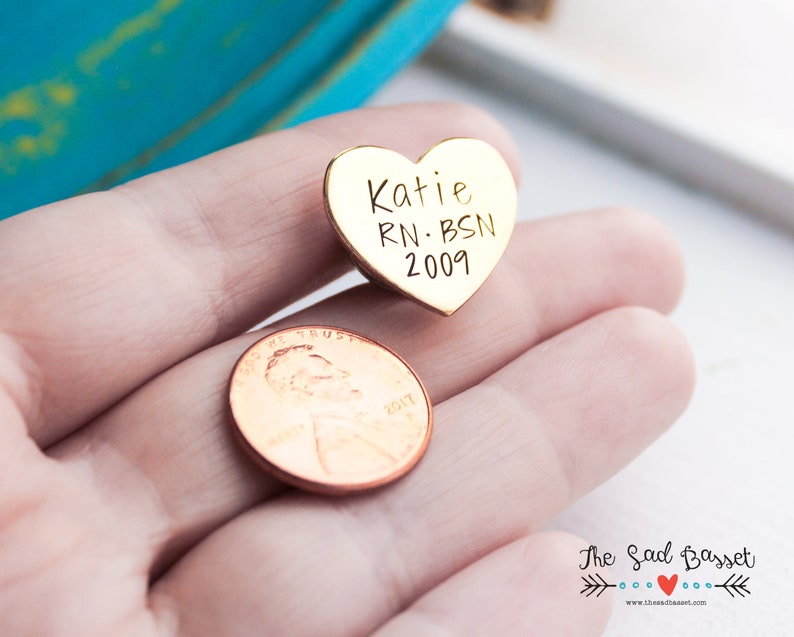 May include: A gold-colored heart-shaped pin with the engraved text "Katie RN BSN 2009". A penny is next to the pin. The pin is likely a personalized gift or keepsake.