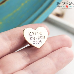 May include: A gold-colored heart-shaped pin with the name "Katie", the initials "RN. BSN", and the year "2009" engraved on it. The pin is held in a person's hand.