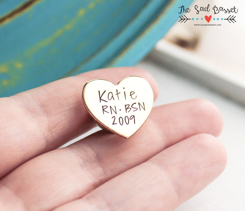 May include: A gold-colored heart-shaped pin with the name "Katie", the initials "RN. BSN", and the year "2009" engraved on it. The pin is held in a person's hand.