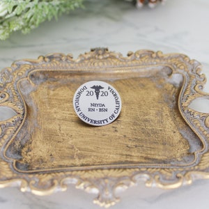 K&ouml;nnte beinhalten: Runder, wei&szlig;-schwarzer Ansteckpin mit dem Text "Dominican University of California" und "2020" mit einem medizinischen Symbol. Der Pin enth&auml;lt auch den Namen "Neyda RN - BSN". Der Pin liegt auf einem goldfarbenen, verzierten Tablett.