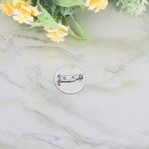 K&ouml;nnte beinhalten: Ein runder, silberfarbener Ansteckbutton mit einem Sicherheitsnadelverschluss auf der R&uuml;ckseite. Der Button befindet sich auf einer wei&szlig;-grauen Marmoroberfl&auml;che, mit gelben Blumen und gr&uuml;nen Bl&auml;ttern im Hintergrund.