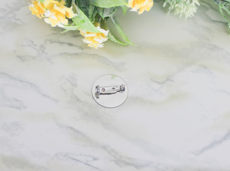 K&ouml;nnte beinhalten: Ein runder, silberfarbener Ansteckbutton mit einem Sicherheitsnadelverschluss auf der R&uuml;ckseite. Der Button befindet sich auf einer wei&szlig;-grauen Marmoroberfl&auml;che, mit gelben Blumen und gr&uuml;nen Bl&auml;ttern im Hintergrund.