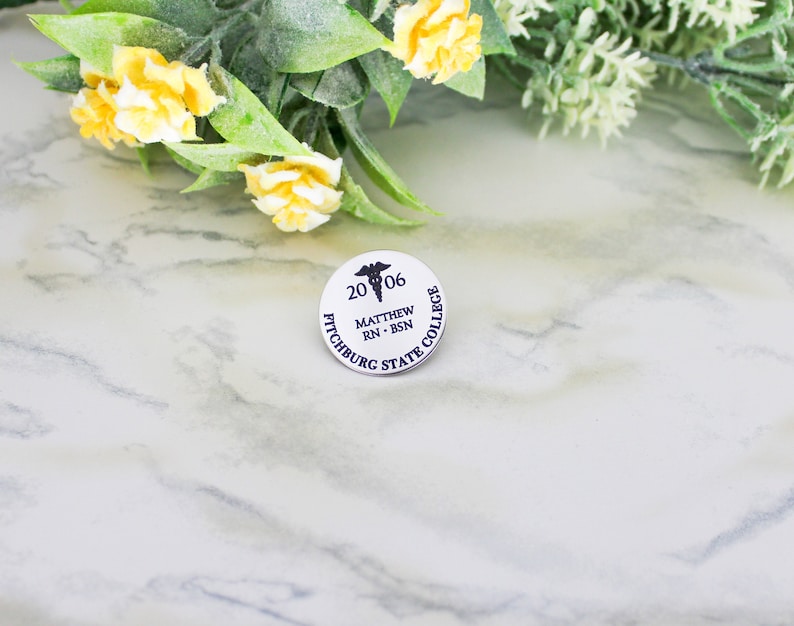 Silver stainless steel round pin with a caduceus at the top center surrounded by the year. Name and credentials below in the center and school name around the edge.