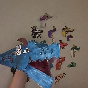 Könnte beinhalten: Eine große blaue Drachen-Handpuppe mit weit geöffnetem Maul, braunen Hörnern und weißen Akzenten. Um die Puppe herum befinden sich mehrere kleinere, bunte Papier-Ausschnitte von verschiedenen Drachenköpfen in Rot, Grün und Violett.