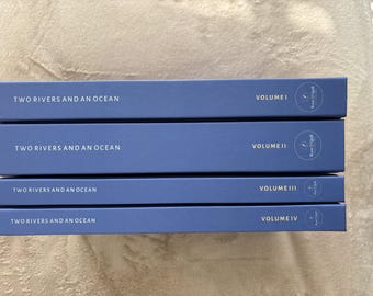 Peut inclure: Une pile de quatre livres bleus intitulés Two Rivers and an Ocean, étiquetés Volume I, Volume II, Volume III et Volume IV. Chaque dos présente un texte blanc et un logo circulaire avec le texte Root & Quill sur le côté droit.