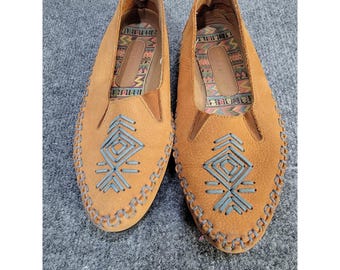 May include: A pair of tan slip-on shoes featuring light blue stitched geometric patterns on the vamps and whip-stitched edges. The interior insoles are lined with a colorful, multi-patterned fabric and include a visible brand logo.