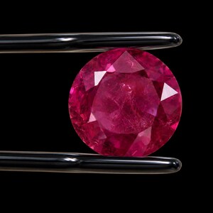 May include: A round, faceted, vibrant pinkish-red gemstone held by a pair of black-tipped metal tweezers. The stone displays a clear, multi-faceted cut that reflects light across its surface, set against a solid, deep black background.