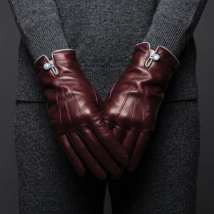 May include: A pair of deep burgundy leather gloves featuring a smooth finish, vertical stitched detailing on the back, and a light grey trim at the wrist. Each glove is secured with a single light grey button closure at the cuff.