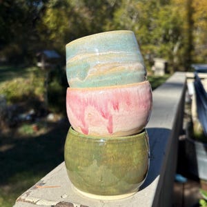 May include: A stack of three ceramic bowls with a rounded shape. The top bowl features a light blue and cream glaze. The middle bowl has a pink glaze with visible drips. The bottom bowl is finished in a solid olive green glaze with a smooth texture.
