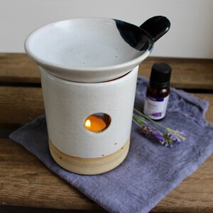May include: A white ceramic essential oil diffuser with a candle inside, a black handle, and a small bottle of oil. The diffuser sits on a wooden surface with lavender sprigs and a purple cloth.