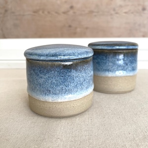 May include: Two ceramic jars with lids. The jars have a cylindrical shape with a light beige base and a blue top that fades to a darker blue. The lids are also blue. The jars are sitting on a beige surface.